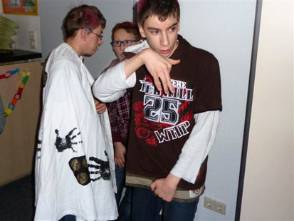 Drei Jungs stehen im Zimmer und unterhalten sich. Einer trägt einen weißen Umhang mit Handabdrücken. 