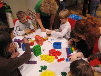 Kinder spielen an einem Tisch mit bunten Bausteinen. Drei Frauen helfen ihnen dabei.