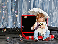 Ein Mädchen sitzt mit Sonnenschirm in einem Koffer. Ringsherum fliegen Seifenblasen.